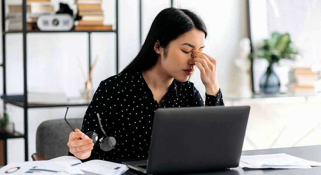 A woman sitting at a desk in front of a computer looking exhausted. She is pinching the bridge of her nose and looks stressed.