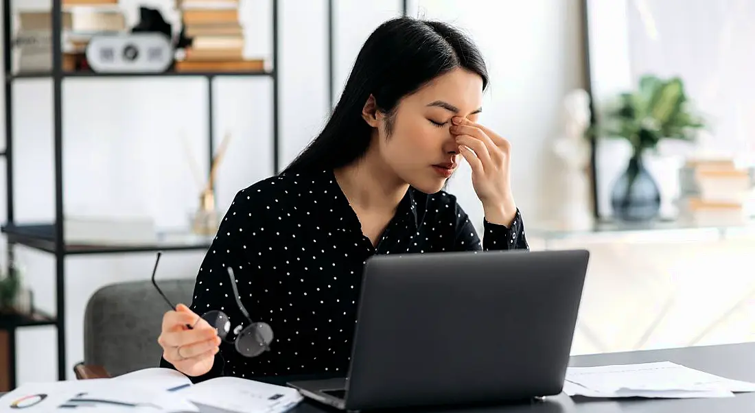 A woman sitting at a desk in front of a computer looking exhausted. She is pinching the bridge of her nose and looks stressed.