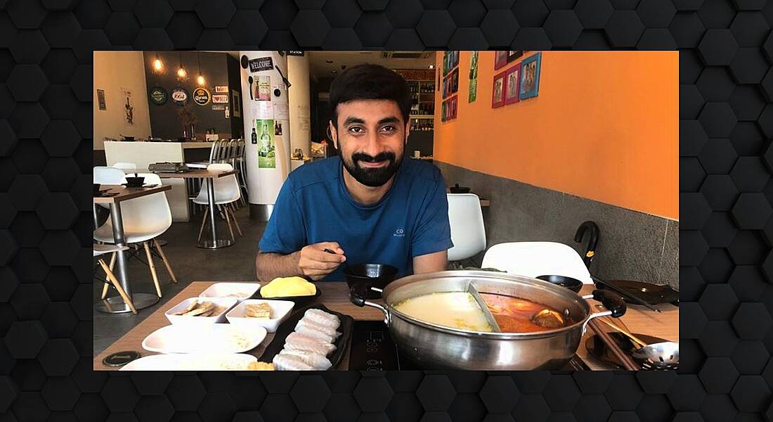 A man, Sachi Gowda, sits at a table in a restaurant with a variety of food in front of him. He is smiling at the camera.