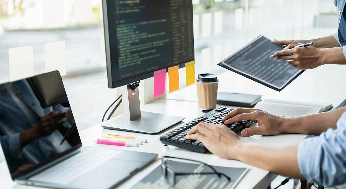 A person types on a keyboard at a large screen that has a lot of code on it. There is a laptop also on the desk.