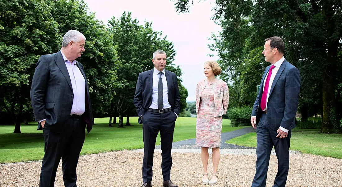 Three men and a women stand socially distanced while having a discussion outside. They are standing on a loose gravel path surrounded by green spaces and trees.