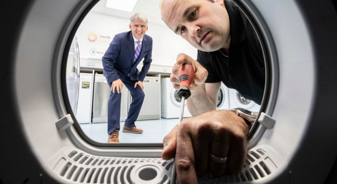 Leo Donovan, CEO of WEEE Ireland stands crouched behind Declan McElwaine, the owner of Electrical Appliance Services as he works on a washing machine with a screwdriver. The image is taken as if from inside the washing machine.