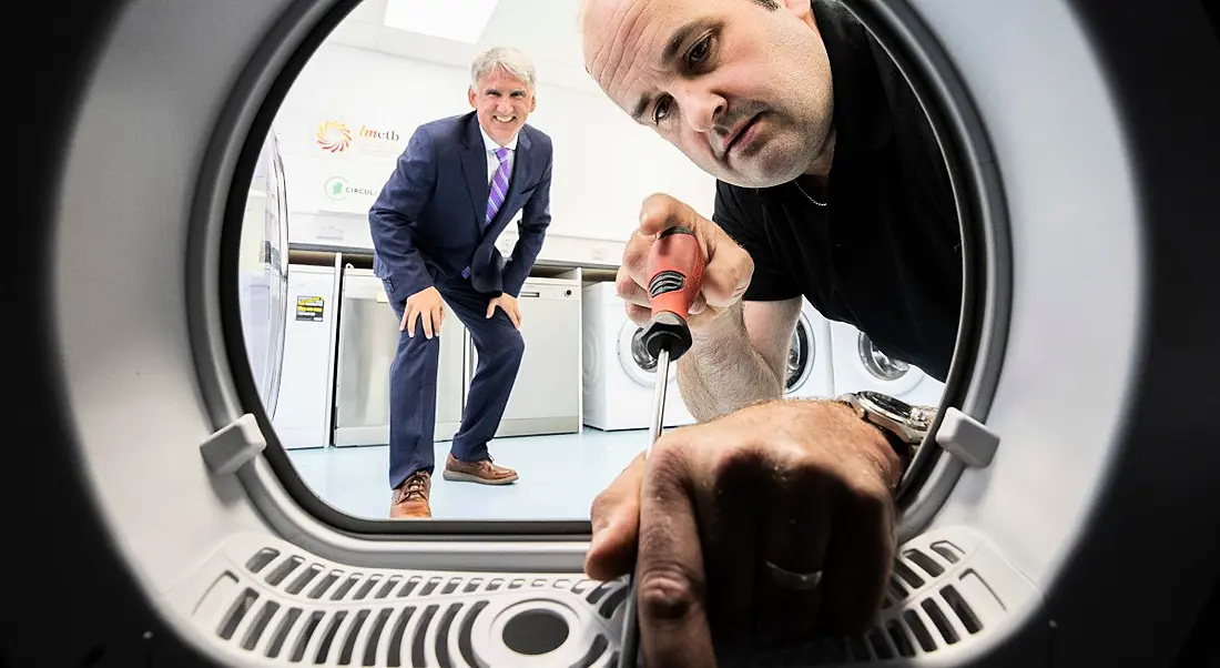 Leo Donovan, CEO of WEEE Ireland stands crouched behind Declan McElwaine, the owner of Electrical Appliance Services as he works on a washing machine with a screwdriver. The image is taken as if from inside the washing machine.