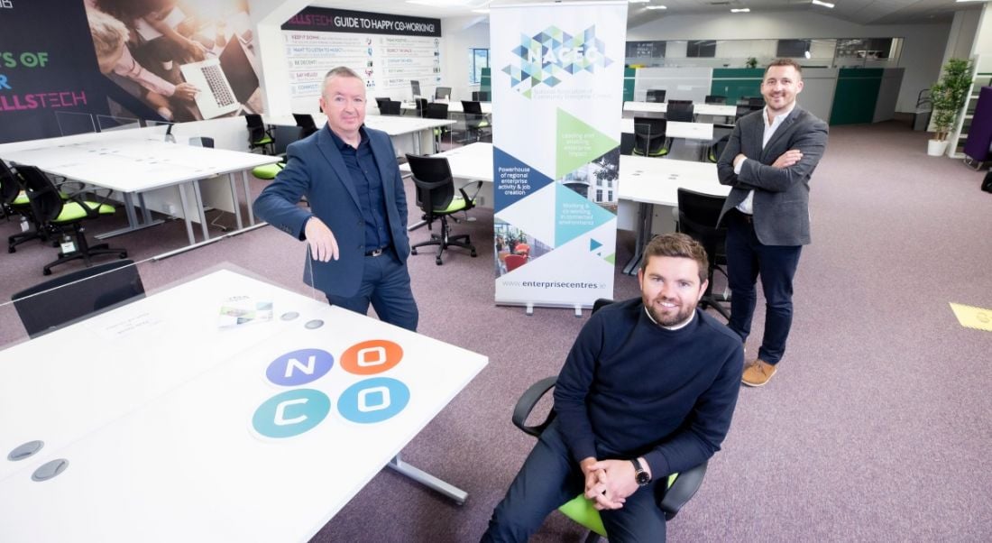 Three men sit in an office space, with the NoCo logo on a desk.