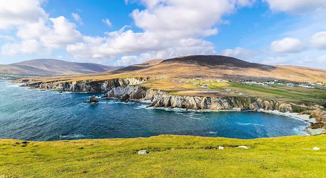 Achill Island is pictured. There is clear blue water on the left, and rolling green hills on the right. There is a blue sky with fluffy clouds interspersed.