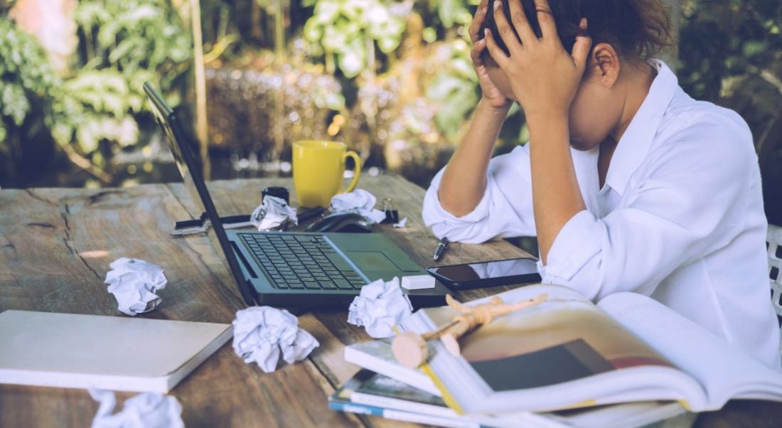 A woman with her head in her hands in front of a laptop at a table covered in scrunched up balls of paper.