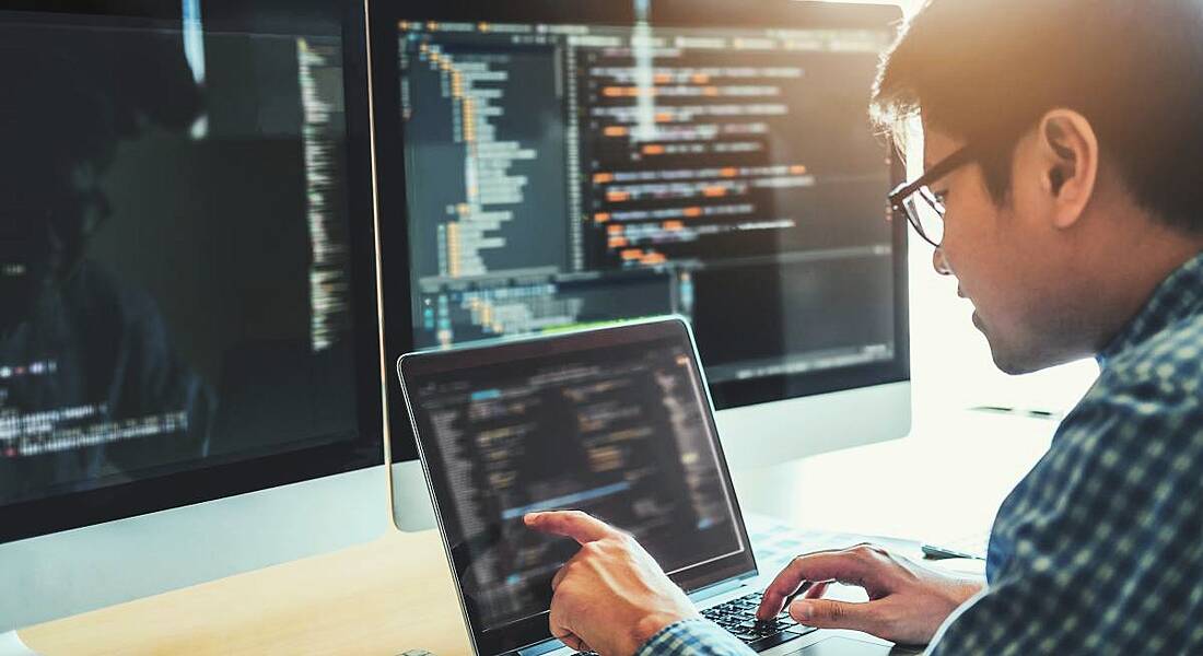 A software developer working on a laptop with two bigger screens on the table. All three screens have lines code on them.