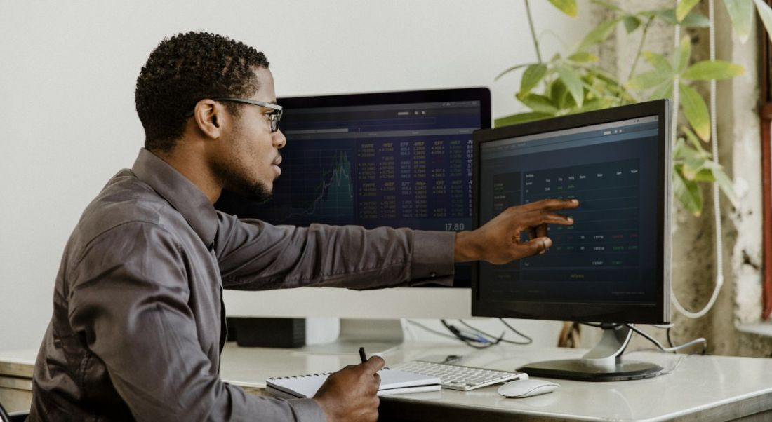 A man working in a brightly lit office at a desk with two screens. He’s pointing at some code on the screen.