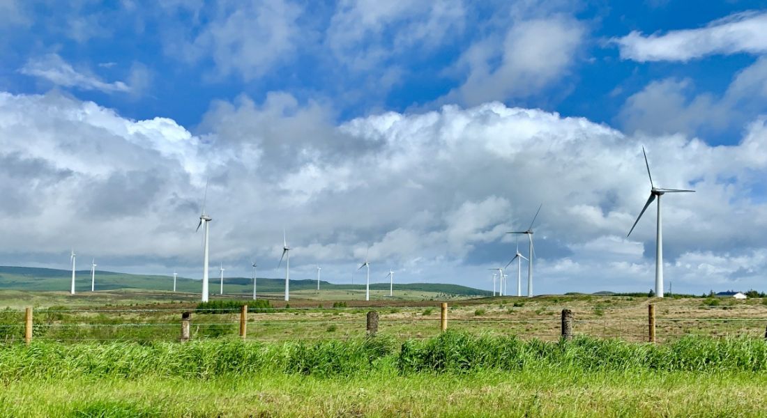 A windfarm in Ireland across a vast green landscape with a fence in the foreground against a blue sky.