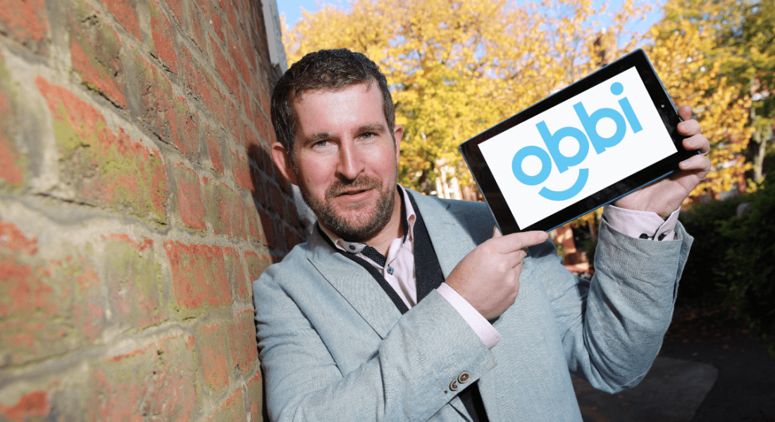 Obbi founder Gareth Macklin leans against a brick wall holding up a tablet device displaying the Obbi logo on its screen.