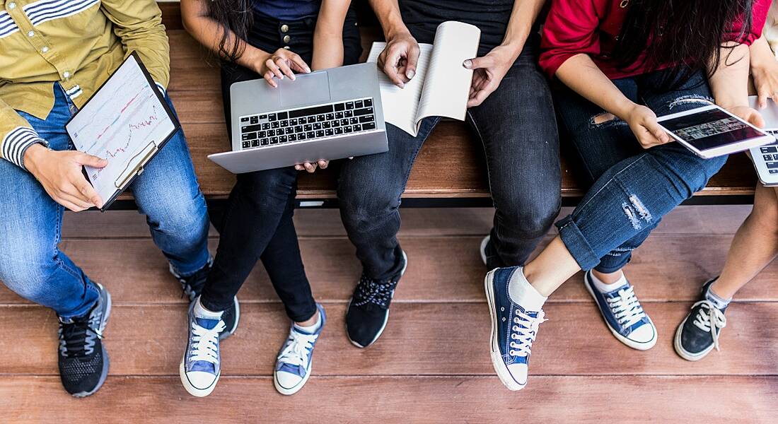 Five college students pictured from the shoulders down, seated on a wooden bench with laptops and notepads.