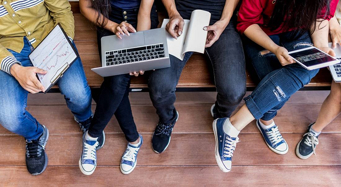 Five college students pictured from the shoulders down, seated on a wooden bench with laptops and notepads.