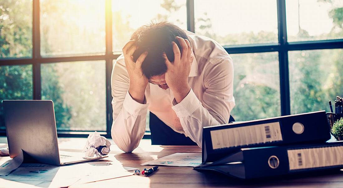 Stressed worker sitting at desk with his head in his hands as light streams in from the window behind him illuminating his workstation.