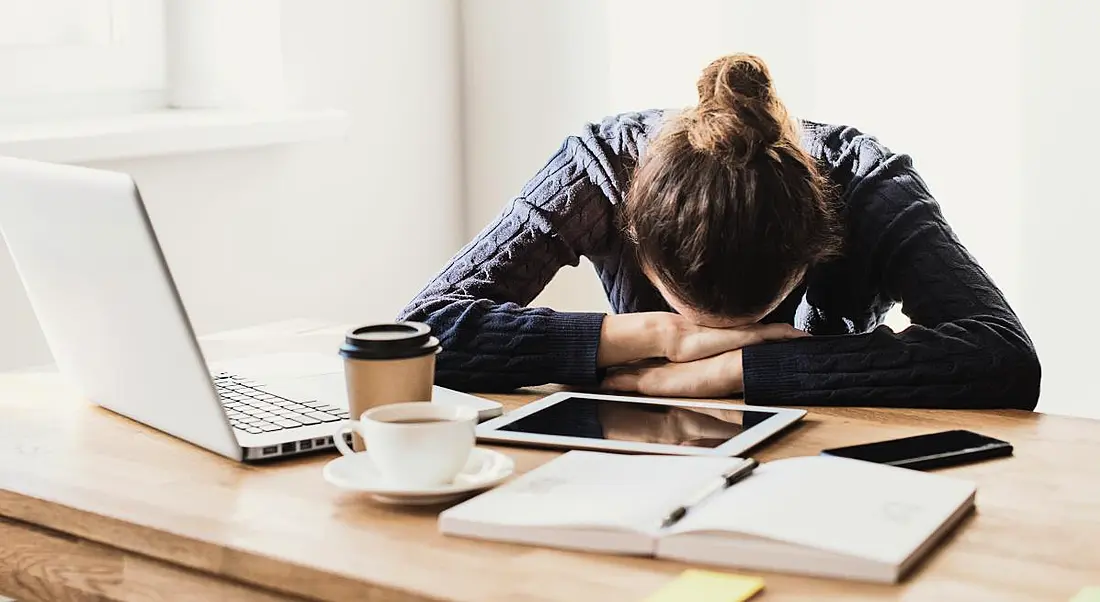 A woman slumped with her head on her arms at a desk. In front of her is a laptop, numerous documents and a coffee cup.