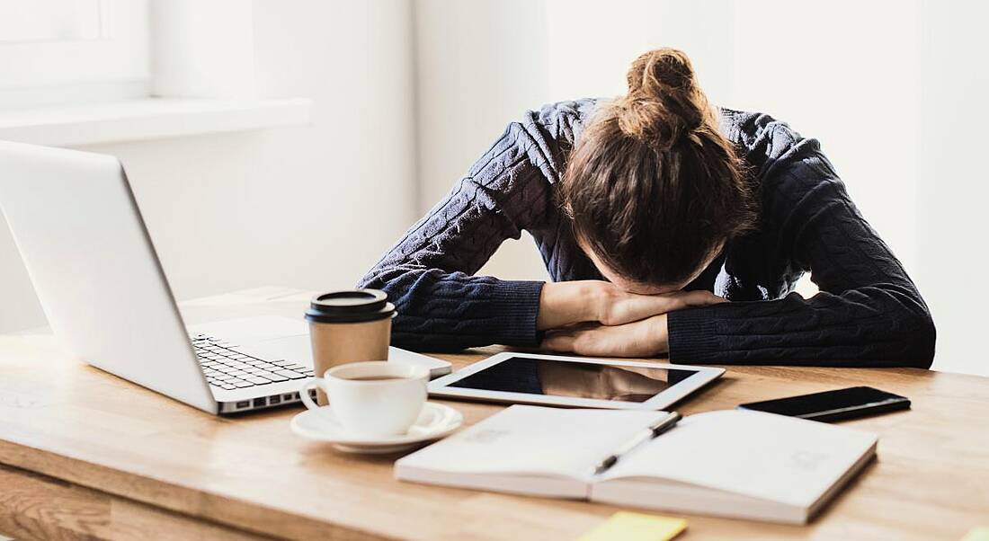 A woman slumped with her head on her arms at a desk. In front of her is a laptop, numerous documents and a coffee cup.