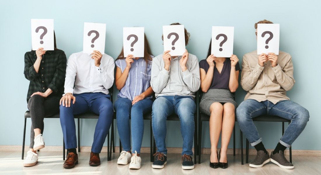 Office workers hiding faces behind paper sheets with question marks while sitting on a row of chairs indoors in an office.