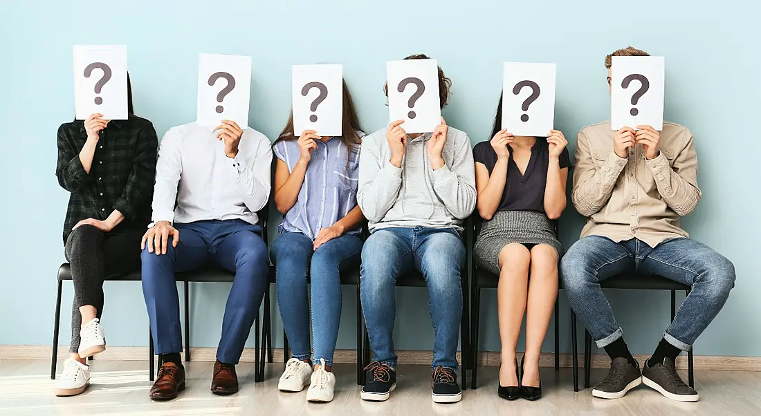 Office workers hiding faces behind paper sheets with question marks while sitting on a row of chairs indoors in an office.