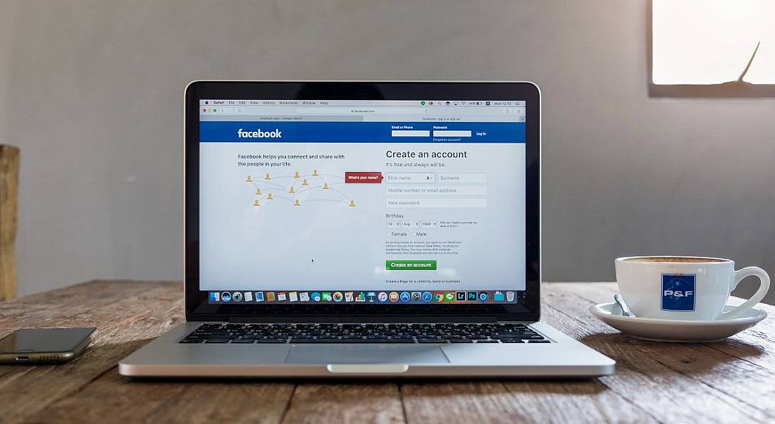 Laptop on a wooden table beside a cup of coffee and a smartphone. Its screen is displaying the login page for Facebook.