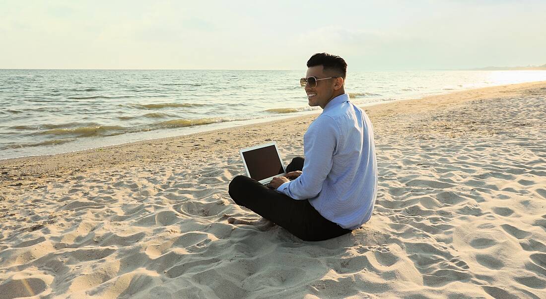 Happy businessman sits cross-legged working with a laptop on the beach.