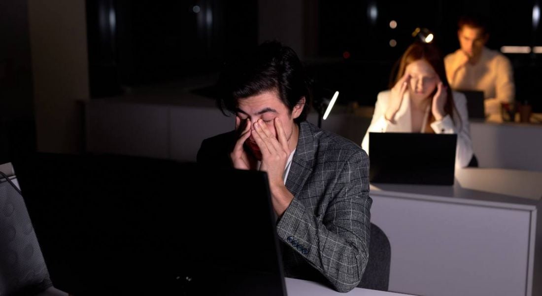 A row of office workers, one behind the other, sit at their desks in front of computers with their heads in their hands. They are tired and working overtime. It is dark outside.