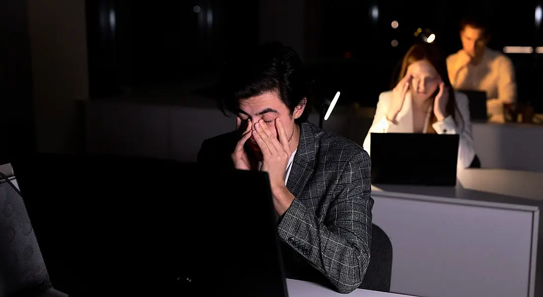 A row of office workers, one behind the other, sit at their desks in front of computers with their heads in their hands. They are tired and working overtime. It is dark outside.