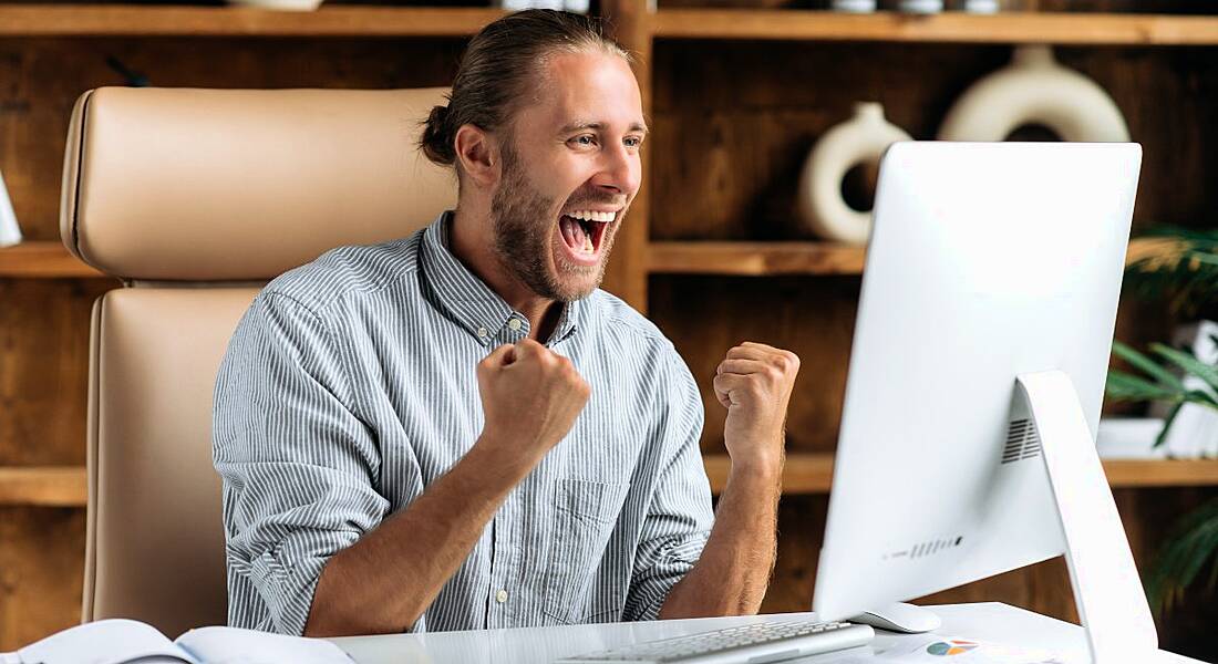 Happy worker sitting at a desk in a modern office rejoicing, raising his fists and yelling at the computer.