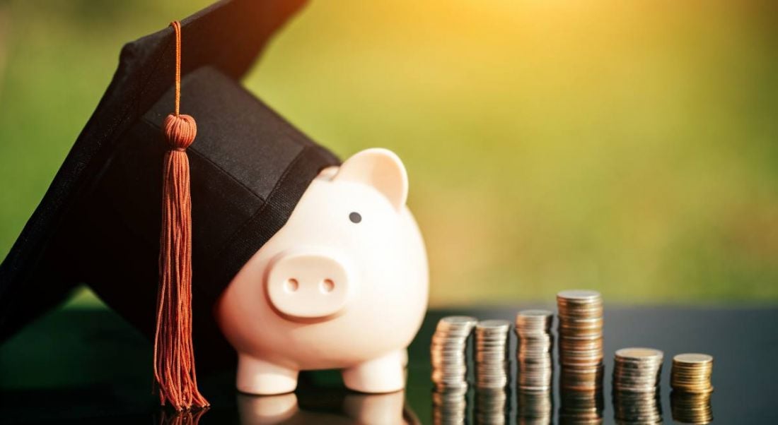 Piggy bank with a graduation cap on it, beside some stacks of coins.