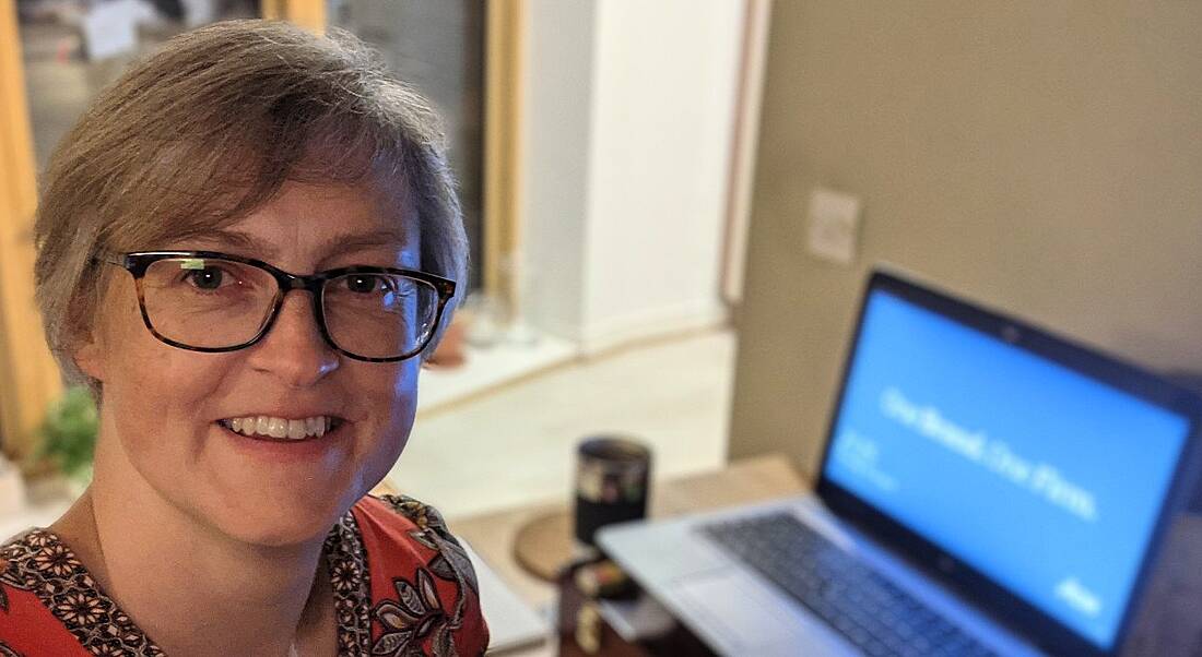 A woman with glasses smiles at the camera while her laptop sits on a desk in the background as she is working from home.