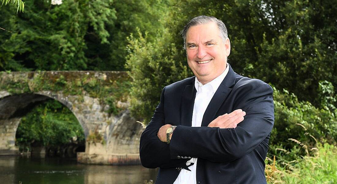 A man in a suit with his arms crossed smiling at the camera. He is standing in an outdoor setting with lots of greenery in the background.