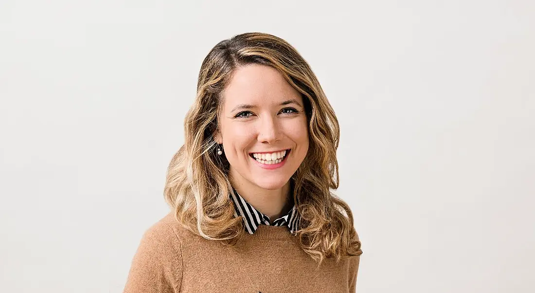 A blonde woman, Dora Palfi, smiles at the camera while standing against a grey background. She is wearing a light brown jumper.