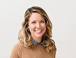 A young woman is remote working at a laptop while holding a phone in a bright co-working space with wooden desks.