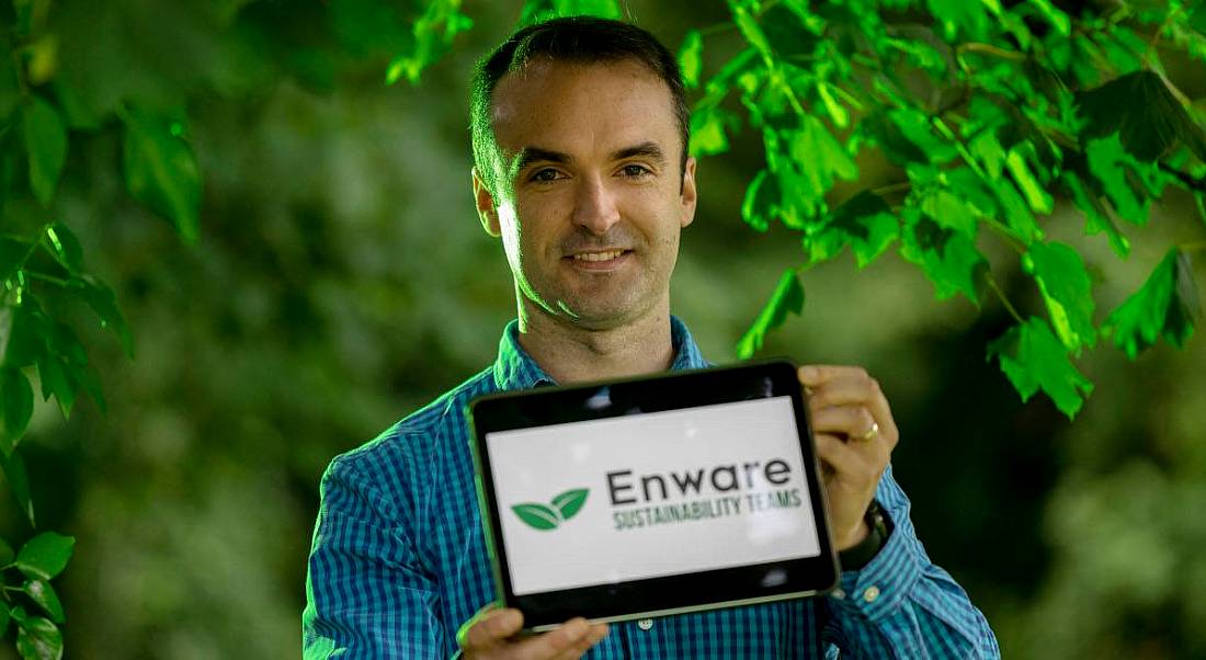 John Keohane pictured holding a tablet device with the Enware logo on the screen, standing against a background of trees with bright green leaves.