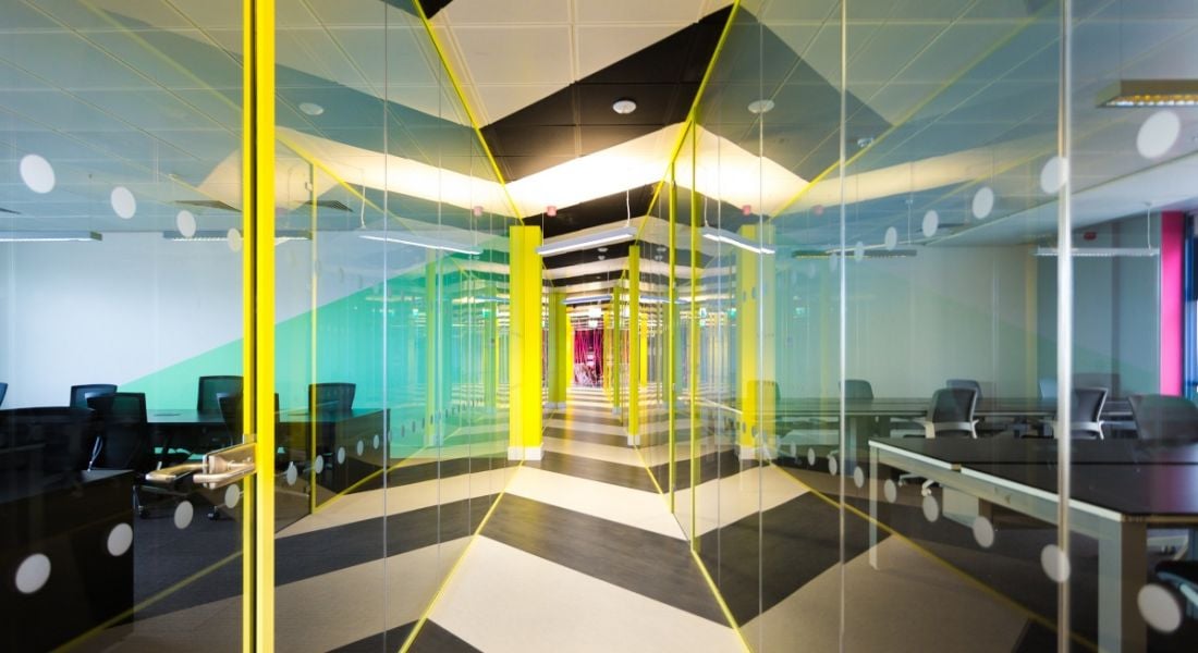 A long glass corridor with yellow lights around the edges. Through the glass is a co-working space for hybrid working.