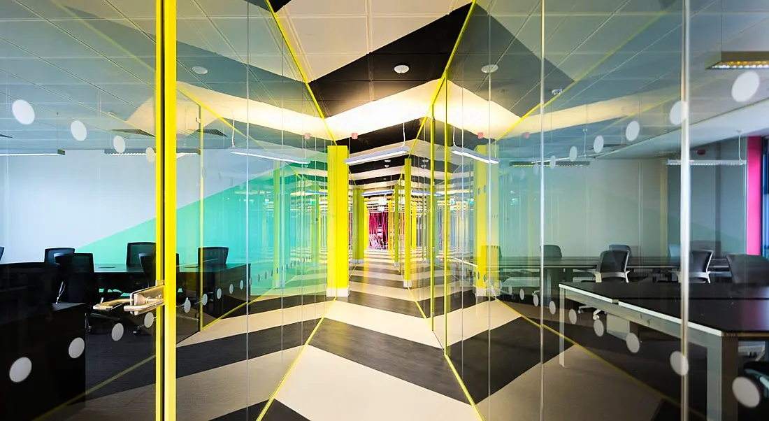 A long glass corridor with yellow lights around the edges. Through the glass is a co-working space for hybrid working.