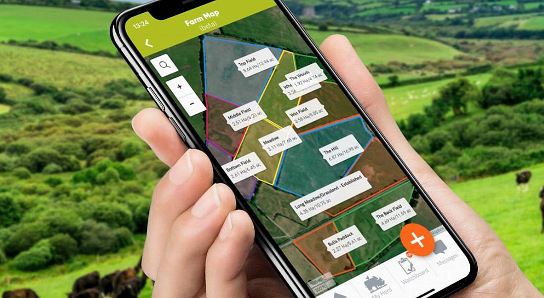 Hand holding a smartphone displaying the Herdwatch farm management app with green fields in the background.