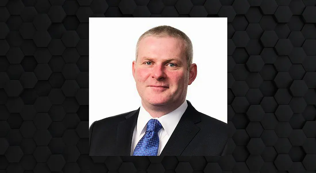 A headshot of Luke Bourke of Avanade in a suit smiling at the camera. The headshot is framed by a black hexagonal design.