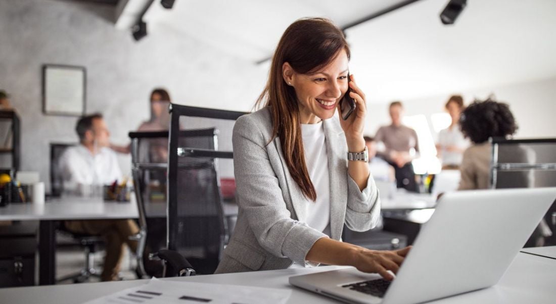 A woman in business attire smiles with a phone up to her ear while working on a laptop. She is leading a team remotely.