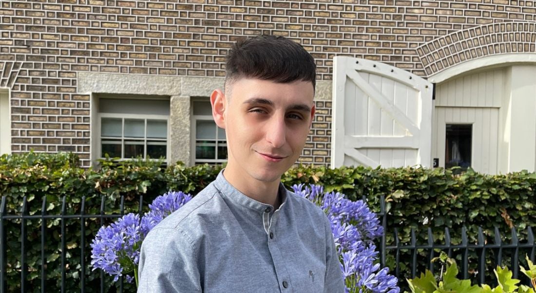 A young man in a blue casual shirt. He is smiling at the camera outdoors. Behind him is a building and several shrubs.