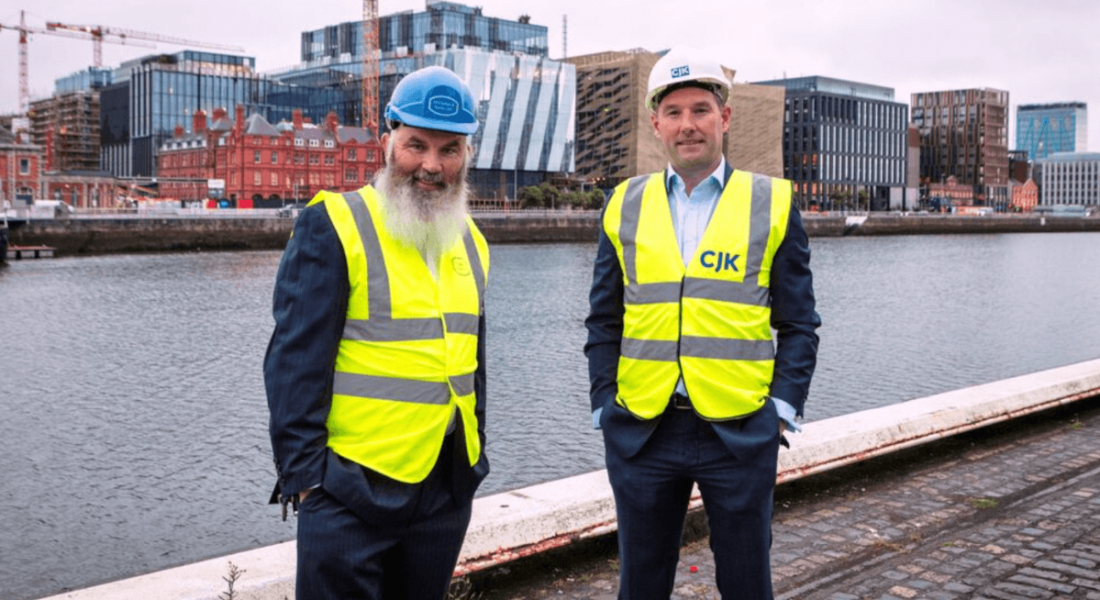 Eamon McGrattanof McGrattan & Kenny and CJK managing director Vinny Bruen stand wearing high-vis clothing on the banks of the river Liffey with buildings and construction behind them.