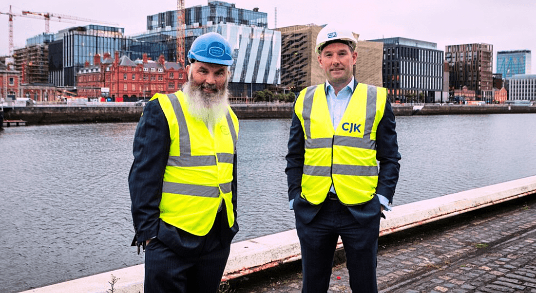 Eamon McGrattanof McGrattan & Kenny and CJK managing director Vinny Bruen stand wearing high-vis clothing on the banks of the river Liffey with buildings and construction behind them.