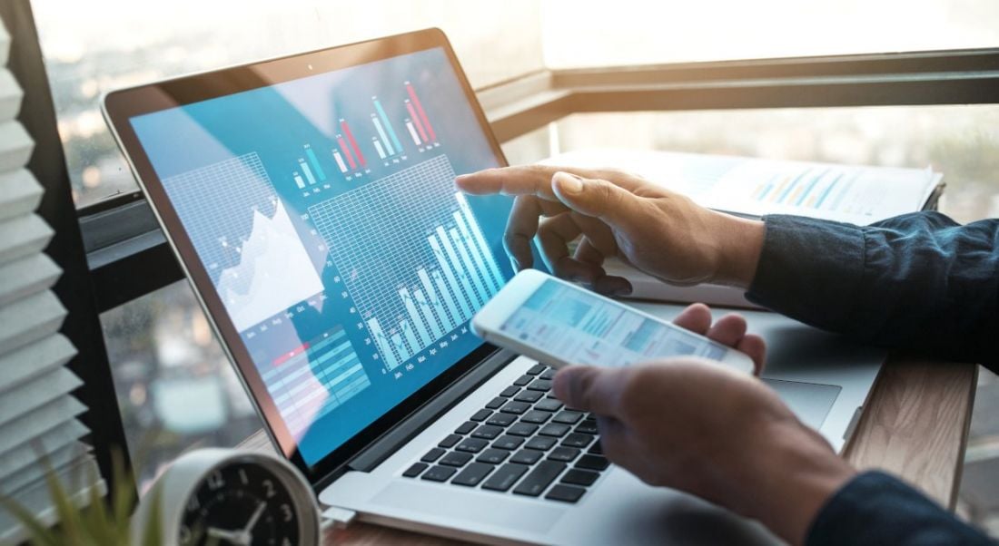 A laptop sitting on a desk with a lot of data on the screen. A person is pointing at it with one hand while looking at more data on their mobile.