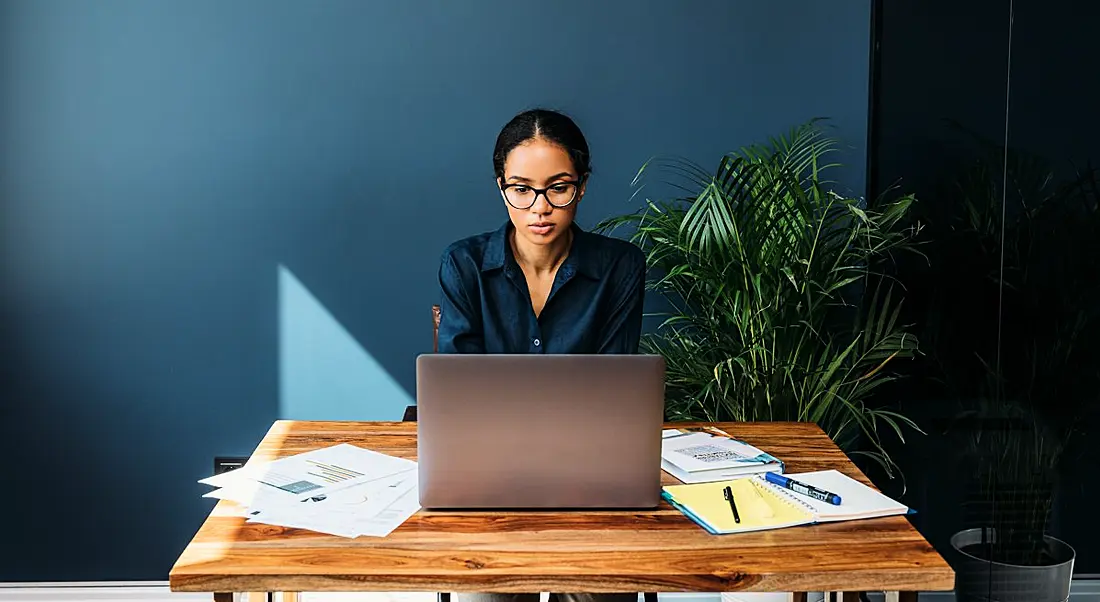 A young woman working on a laptop at a wooden desk surrounded by sheets of paper. There is a plant behind her against a navy wall.