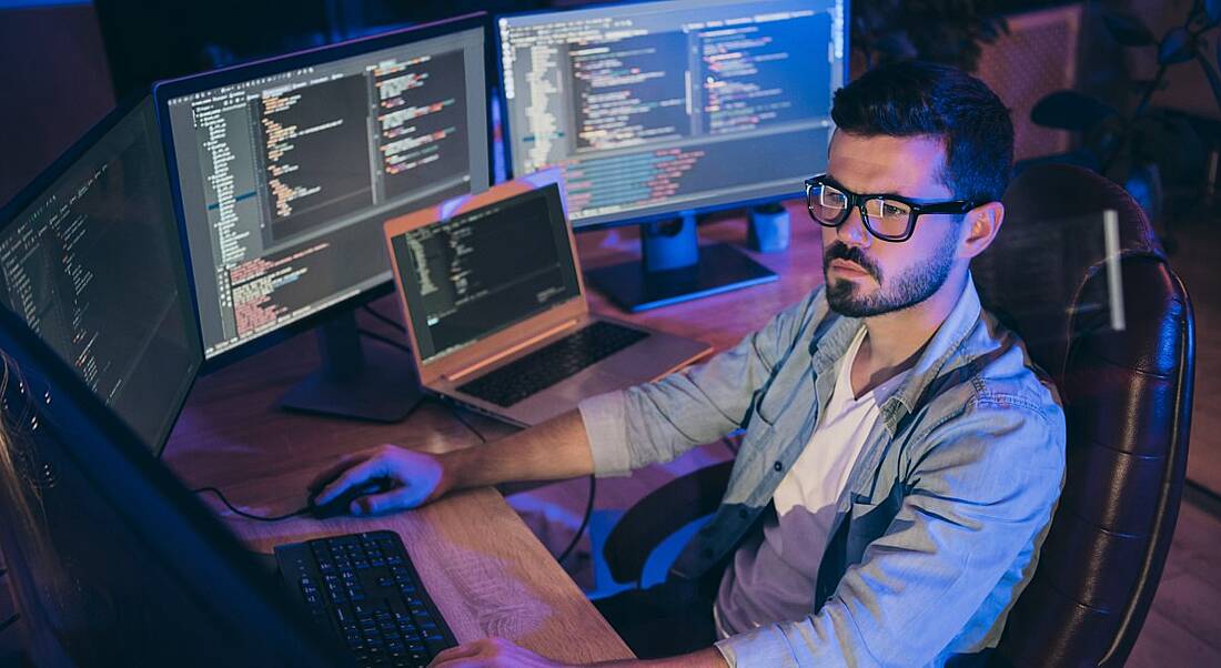 A male tech worker in causal clothing sits at a corner desk working at several monitors, symbolising a remote workforce.