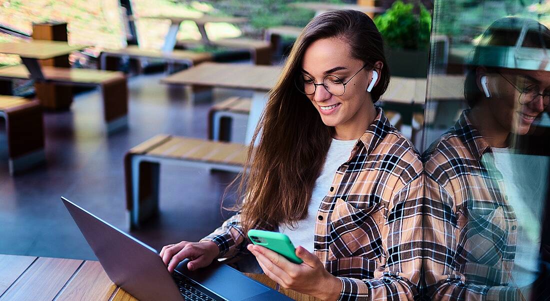 A young woman is remote working at a laptop while holding a phone in a bright co-working space with wooden desks.