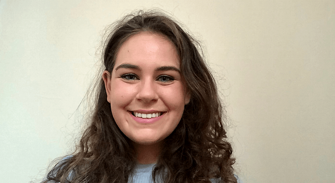 A young smiling woman with curly brown hair stands against a plain wall. She is a graduate working at Avanade.