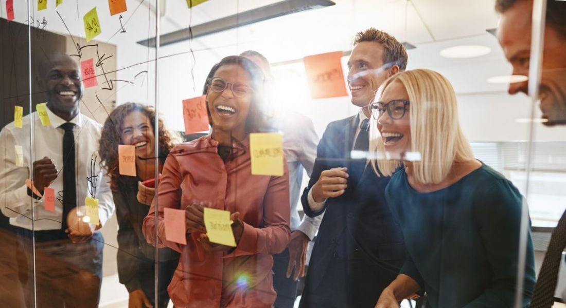 A group of happy business people having a very fun brainstorming session in an office with a glass wall and post-it notes all over it.