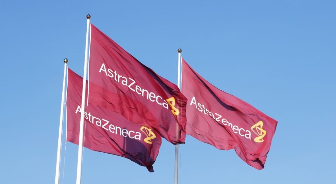 Three purple flags with the logo for Atrazeneca flying in the wind on top of flagpoles, against a clear blue sky.