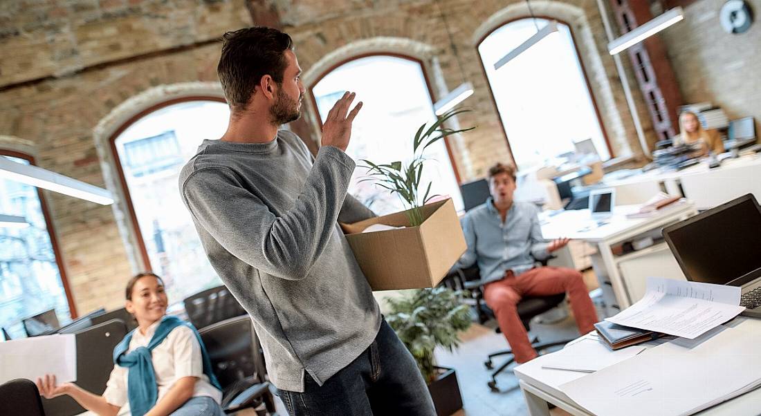 Man waving goodbye to shocked colleagues in a bright airy office as he executes his great resignation, carrying a cardboard box.