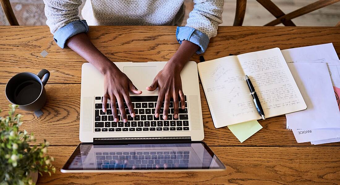 Woman's hands on laptop computer keys as she works remotely or from home at a table covered in papers.