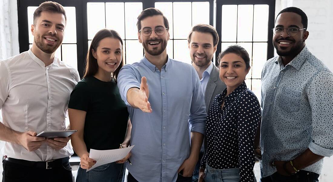 Workers/ friendly HR department stand in a group smiling while one of them outstretches his hand in welcoming gesture.